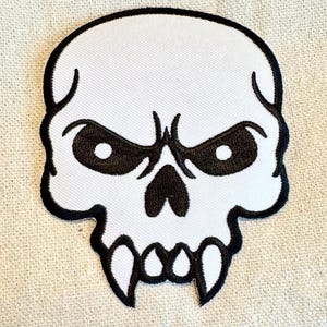 May include: White embroidered skull patch with black eyes and fangs. The skull has a menacing expression.