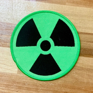 Puede incluir: Un parche bordado verde y negro con un símbolo de radiación. El símbolo es un trébol de tres hojas estilizado con un círculo negro en el centro.