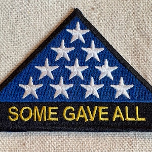 May include: A triangular patch featuring a blue field with white stars, resembling a folded American flag. The bottom section is black with the embroidered text "SOME GAVE ALL" in gold. The patch has a black border.