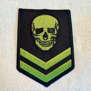 May include: A black and green embroidered patch with a skull and crossbones design. The skull is green and the crossbones are black. The patch has two green stripes.