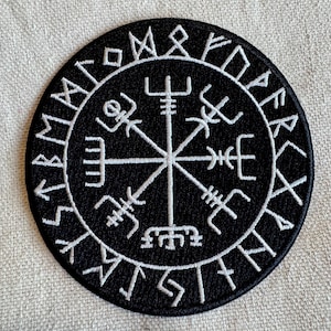 May include: A black and white embroidered patch with a compass design and runes around the edge. The compass has a central point with four arms extending out to the edges of the circle. The runes are in a circle around the compass.