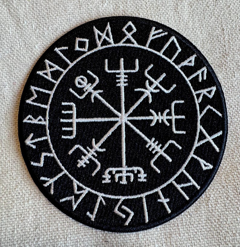 May include: A black and white embroidered patch with a compass design and runes around the edge. The compass has a central point with four arms extending out to the edges of the circle. The runes are in a circle around the compass.