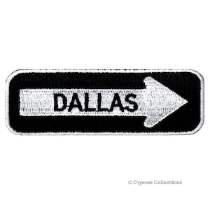 May include: A black and white embroidered patch with the word "DALLAS" and a white arrow pointing right. The patch has a rectangular shape with rounded corners and a white border.