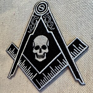 May include: Black and white embroidered patch of a skull inside a square and compass, a symbol of Freemasonry.