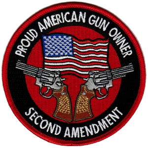 Puede incluir: Una bandera estadounidense roja, blanca y azul con dos revólveres plateados cruzados detrás de ella. La bandera está rodeada por un círculo negro con el texto "Proud American Gun Owner Second Amendment" en blanco.