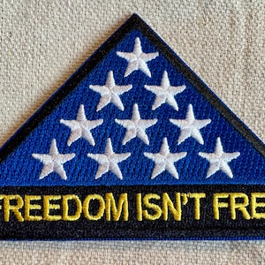 Op de afbeelding: Een driehoekige patch met een blauwe achtergrond en witte sterren. Het onderste gedeelte is zwart met de geborduurde tekst "FREEDOM ISN'T FREE" in geel. De patch heeft een zwarte rand.