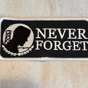 Pode incluir: Um remendo bordado preto e branco com uma silhueta de uma pessoa em frente a uma torre e arame farpado. O texto "NEVER FORGET" é bordado em branco sobre um fundo preto.