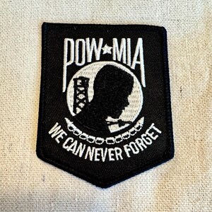Può includere: Toppa ricamata nera con scritta bianca "POW MIA WE CAN NEVER FORGET". La toppa presenta una silhouette bianca di una testa umana con una catena attorno al collo.