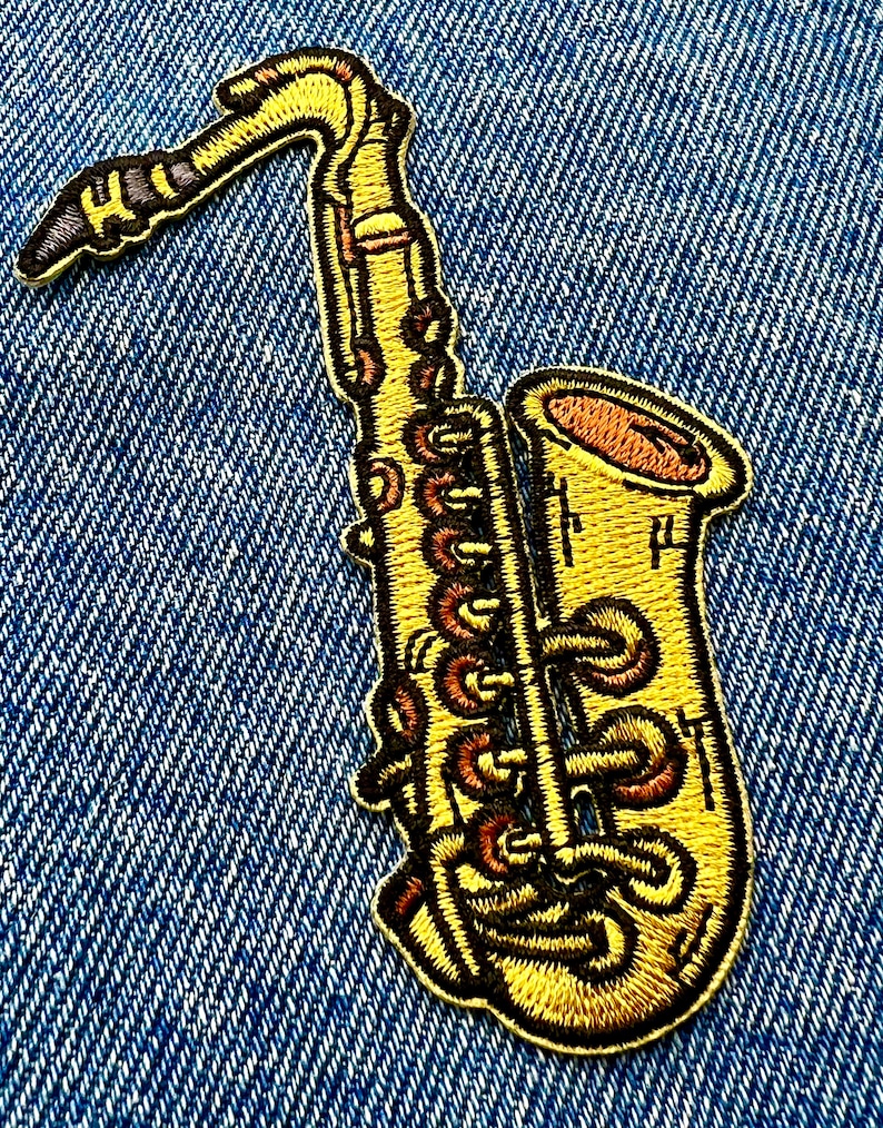 May include: A yellow embroidered saxophone patch with brown details. The patch is on a blue denim background.