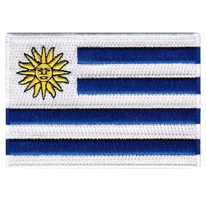 Pode incluir: Uma bandeira branca com nove faixas azuis horizontais e um sol amarelo com rosto no canto superior esquerdo. A bandeira é feita de tecido e tem uma superfície ligeiramente texturizada.