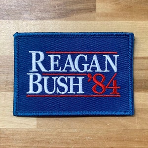 May include: A blue fabric patch with white and red embroidery. The text on the patch reads "REAGAN BUSH '84".