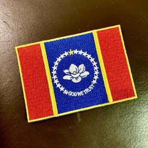 Pode incluir: Um remendo bordado azul, vermelho e amarelo com uma flor de magnólia branca no centro. O remendo tem uma borda amarela e as palavras "In God We Trust" estão escritas em branco.