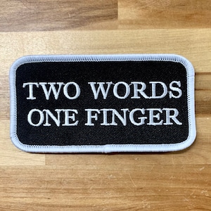 May include: Black and white embroidered patch with white stitching around the edges. The patch says "TWO WORDS ONE FINGER" in white text.