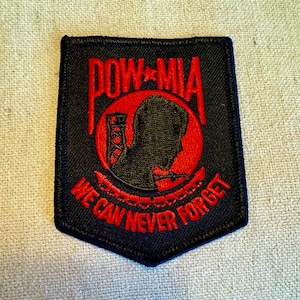 Może przedstawiać: Czarna naszywka haftowana z czerwonym napisem "POW-MIA WE CAN NEVER FORGET" i czerwonym kółkiem z sylwetką głowy żołnierza w środku.