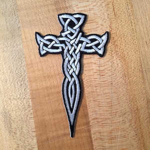 May include: A silver embroidered Celtic cross patch with a pointed bottom. The cross is made of interwoven knots.