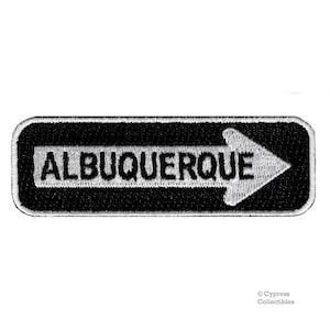 Könnte beinhalten: Schwarzer und weißer gestickter Aufnäher mit einem weißen Pfeil, der nach rechts zeigt, und dem Text "ALBUQUERQUE" in weißen Buchstaben.