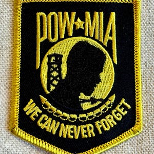 May include: A black and yellow embroidered patch with the words "POW-MIA" and "We Can Never Forget". The patch features a silhouette of a prisoner of war.