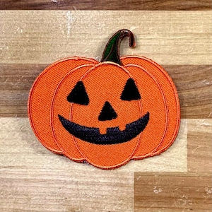 May include: Orange felt pumpkin with a black smiling jack-o'-lantern face. The pumpkin is on a wooden background.