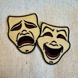 May include: Two embroidered fabric patches depicting theatrical masks. One mask is a traditional comedy mask with a wide smile and red lips. The other mask is a traditional tragedy mask with a frown and red lips.