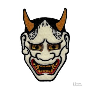 May include: A white embroidered Japanese demon mask with red and gold details. The mask has horns, a wide mouth, and large eyes.