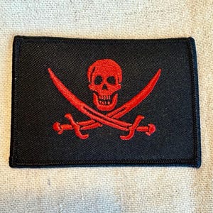 Może przedstawiać: Czarna łatka z tkaniny z haftowanym na czerwono motywem czaszki i skrzyżowanych kości. Czaszka ma szczegółowy wzór z wyraźnym nosem i zębami.