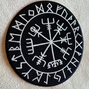 May include: Black embroidered patch with a white compass design and runes around the edge. The compass has four points with symbols at each point. The runes are in a circle around the compass.
