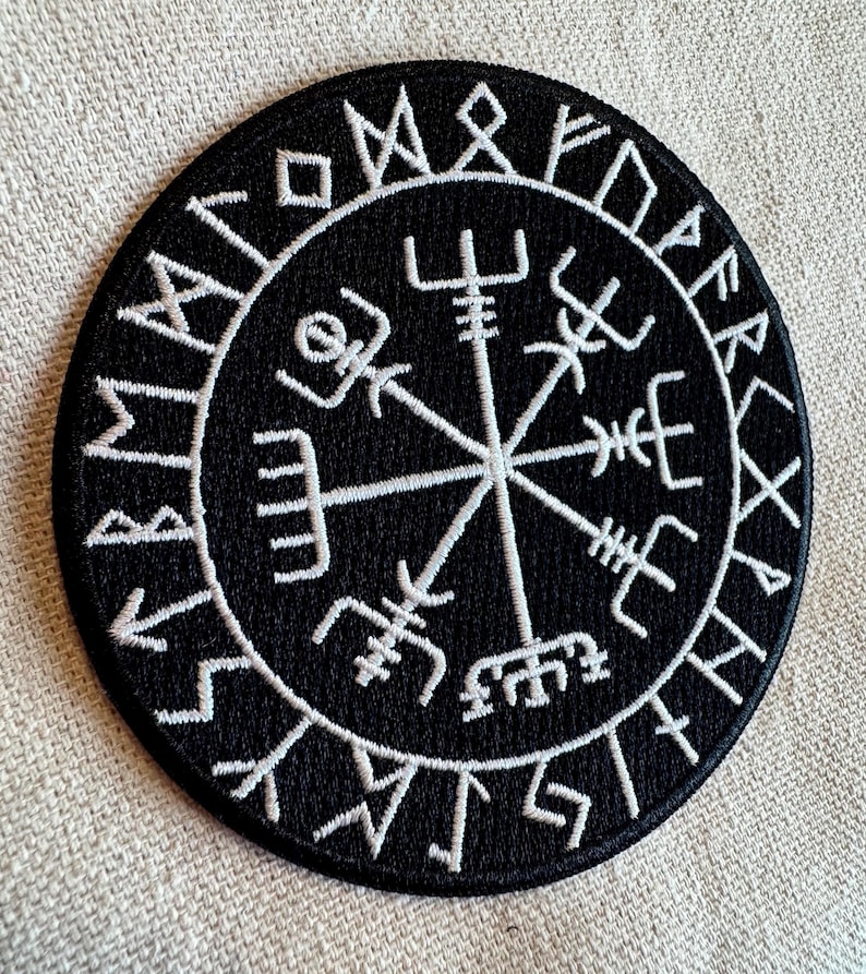 May include: Black embroidered patch with a white compass design and runes around the edge. The compass has four points with symbols at each point. The runes are in a circle around the compass.