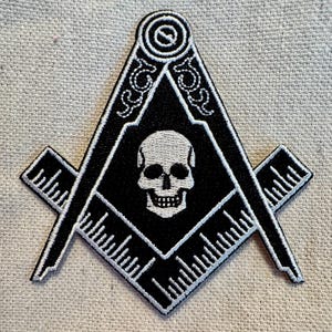 Könnte beinhalten: Schwarz-weiß gestickter Aufnäher mit einem Schädel in einem Quadrat und Zirkel, einem traditionellen Symbol der Freimaurerei.