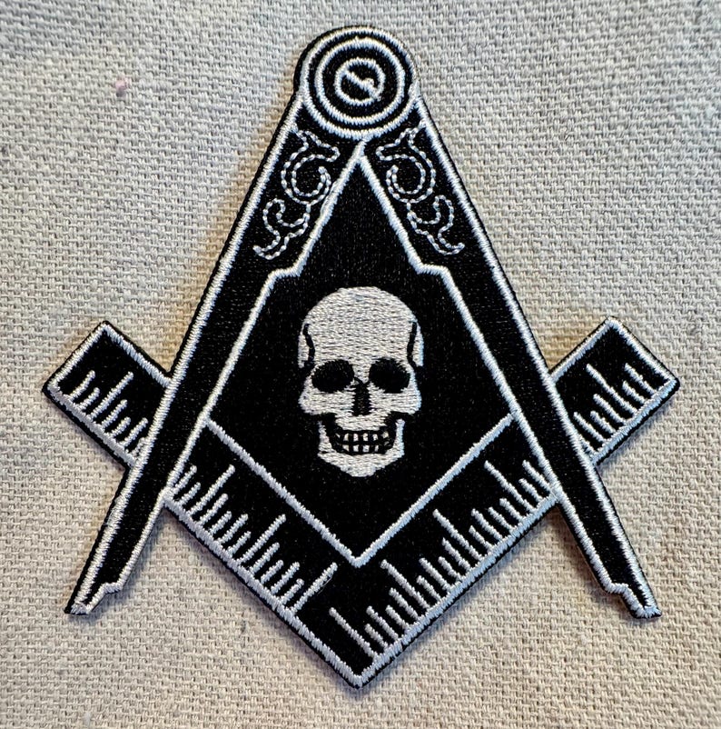 May include: Black and white embroidered patch of a skull inside a square and compass, a traditional symbol of Freemasonry.