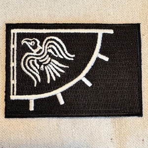May include: Black embroidered patch featuring a white stylised raven design. The raven is facing left with its wings spread. The patch has a white border and a white semi-circle with white spikes at the bottom.