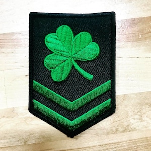 May include: A black and green embroidered patch with a four-leaf clover and two green chevron stripes.