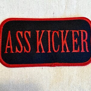 May include: Black fabric patch with red stitching and the text "ASS KICKER" in red embroidered letters.