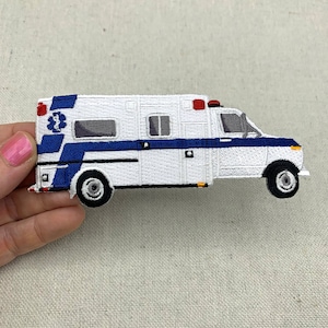 May include: White and blue embroidered patch of an ambulance with a red light on top. The ambulance has a blue stripe with a white star of life symbol on the side.