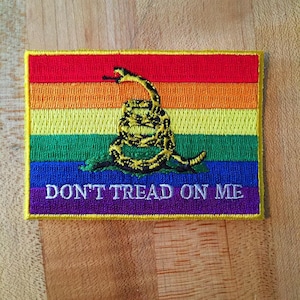 Può includere: Una toppa color arcobaleno con un serpente giallo avvolto attorno a un bastone e le parole "Don't Tread On Me" in bianco.