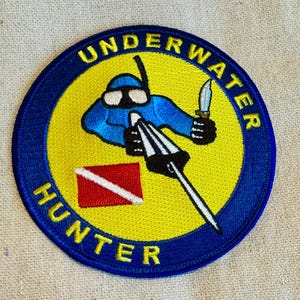 Può includere: Una toppa ricamata blu e gialla con il testo "UNDERWATER HUNTER" in un cerchio blu. Un subacqueo dei cartoni animati con un fucile subacqueo e un coltello è al centro della toppa.