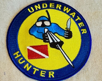 Może przedstawiać: Naszywka haftowana w kolorze niebieskim i żółtym z tekstem "UNDERWATER HUNTER" w niebieskim kółku. W środku naszywki znajduje się kreskówkowy nur z włócznią i nożem.