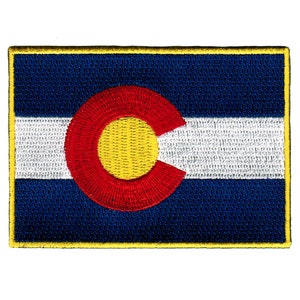 Pode incluir: Etiqueta bordada da bandeira do estado do Colorado. A bandeira é azul com uma faixa horizontal branca no centro. Um grande círculo vermelho com um centro amarelo está no centro da bandeira.