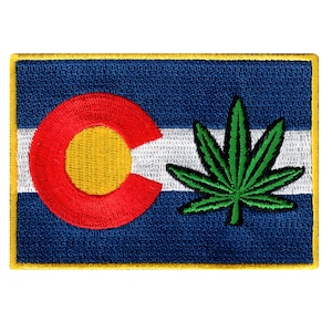 May include: A blue and white Colorado state flag with a green marijuana leaf embroidered on the right side.