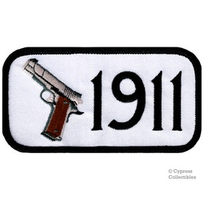 Puede incluir: Un parche rectangular blanco con un borde negro. El parche presenta una ilustración en blanco y negro de una pistola con el número "1911" en negro.