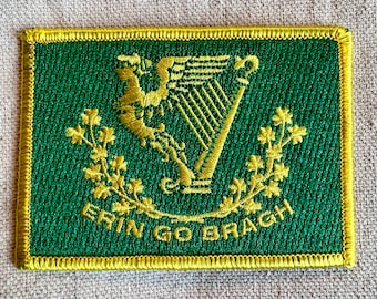 Può includere: Una toppa ricamata verde con un bordo dorato. La toppa presenta un'arpa dorata con un motivo a trifoglio dorato su entrambi i lati. Il testo "Erin Go Bragh" è ricamato in oro nella parte inferiore della toppa.