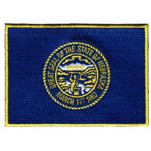 May include: A blue embroidered patch with a yellow border. The patch features the Great Seal of the State of Nebraska, which depicts a depiction of a river, a bridge, and a rising sun. The text "Great Seal of the State of Nebraska" and "March 1st 1867" are visible on the seal.