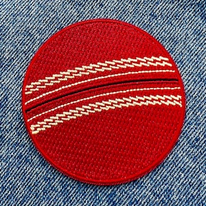 May include: Red embroidered cricket ball patch with white stitching. The ball has a textured surface and a classic design.