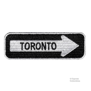 May include: Black and white embroidered patch with a white arrow pointing to the right and the word "TORONTO" in black.