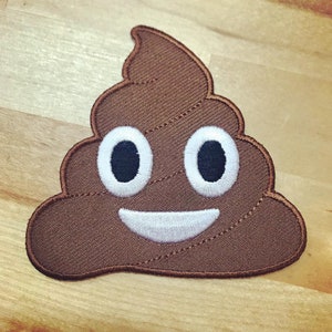 May include: A brown felt patch with a smiling poop emoji. The emoji has white eyes and a white mouth.