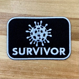 Może przedstawiać: Czarna prostokątna naszywka z białym haftowanym tekstem i grafiką wirusa. Słowo "SURVIVOR" jest wyszyte na biało pod stylizowanym wizerunkiem wirusa. Naszywka ma białą obwódkę.