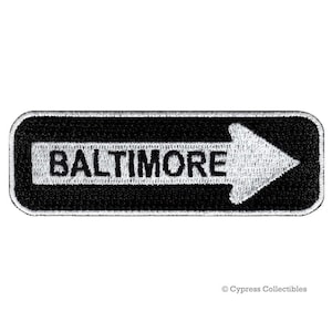 May include: Black and white embroidered patch with a white arrow pointing to the right and the word "BALTIMORE" in black letters.