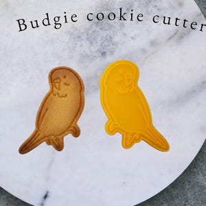 Puede incluir: Una galleta horneada y un cortador de galletas de plástico amarillo, ambos con forma de periquito, están sobre una superficie de mármol. La galleta es de color marrón claro, mientras que el cortador es de color amarillo brillante. El texto superior dice "Budgie cookie cutter" y "typeB" en la parte inferior.