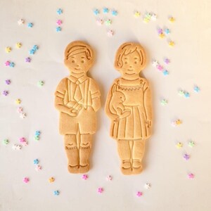 以下が含まれることがあります： 男の子と女の子の形をした2つのジンジャーブレッドクッキーが並んでいます。男の子のクッキーは、ショートパンツ、シャツ、ネクタイを着ています。女の子のクッキーはドレスを着て、ウサギを持っています。カラフルな星型のスプリンクルが白い表面に散らばっています。