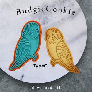 Puede incluir: Dos cortadores de galletas con forma de periquito. Uno es turquesa con un contorno naranja, el otro es una galleta de color marrón dorado. También se ve el texto "BudgieCookie", "TypeC" y "download stl".