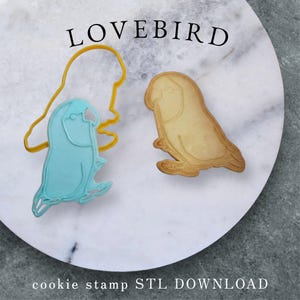 Puede incluir: Un cortador de galletas azul con forma de ave del amor junto a una galleta horneada y un contorno amarillo del cortador. La palabra "LOVEBIRD" está arriba y "cookie stamp STL DOWNLOAD" abajo. Sobre una superficie de mármol blanco.